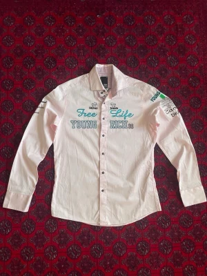 Skjorta Young & Rich - Säljer en ljusrosa skjorta från Young & Rich med broderade detaljer och texten 'Free Life', 'Cayman Islands' och 'Young Rich 96' framtill. Skjortan har lång ärm, knappar och kontrastfoder i kragen. Perfekt för dig som gillar unika statement-plagg. Riktig fet unik skjorta inför sommaren. 