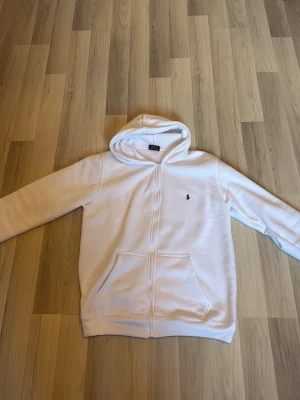 Vit hoodie från Polo Ralph Lauren - Snygg vit hoodie med dragkedja från Polo Ralph Lauren. Klassisk design med huva, två fickor framtill och den ikoniska broderade loggan på bröstet. Perfekt för en clean och stilren look. Mjuk bomullskänsla och relaxed fit.