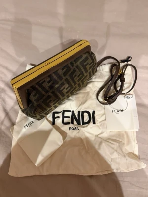 Fendi brun kuvertväska med FF-mönster - Snygg kuvertväska från Fendi i brunt tyg med klassiskt FF-logomönster. Väskan har guldfärgade metalldetaljer och en avtagbar axelrem i läder. Perfekt för dig som vill ha en ikonisk accessoar med lyxig känsla.
