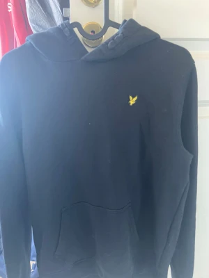 Svart hoodie från Lyle & Scott - Svart hoodie från Lyle & Scott med klassisk gul logga på bröstet. Hoodien har huva med dragsko och en stor magficka framtill. Tillverkad i mjukt material, perfekt för en chill och avslappnad stil.