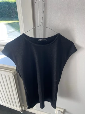 Svart ribbad topp från Zara - En svart ribbad topp från Zara med rund halsringning och ärmlös design. Toppen har en enkel och stilren look som passar perfekt till jeans eller kjol. Materialet är mjukt och stretchigt för en bekväm passform.