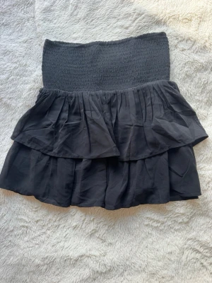 Svart volangkjol - Supersnygg svart kjol med bred smock i midjan och dubbla volanger. Kjolen har en kort modell och är perfekt att styla med en enkel topp eller hoodie. Materialet känns lätt och flowy.