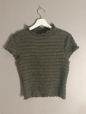 Olivgrön ribbad topp med kort ärm - Trendig olivgrön topp med ribbad struktur och korta ärmar. Toppen har en lätt croppad passform och hög halsringning. Perfekt att styla med jeans eller kjol för en cool vardagslook.