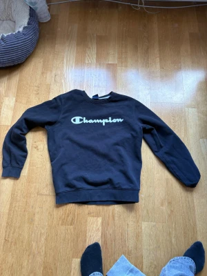 Svart Champion sweatshirt - Svart sweatshirt från Champion med klassisk logga i vitt över bröstet. Tröjan har rund halsringning, långa ärmar och ribbade muddar vid ärmslut och nederkant. Perfekt för en avslappnad och sportig stil.