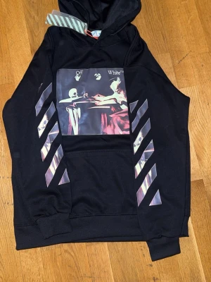 Svart Off-White hoodie med tryck - Svart hoodie från Off-White med stort grafiskt tryck på bröstet och diagonala ränder på ärmarna. Klassisk huva och magficka. Materialet är mjukt och bekvämt, perfekt för en streetwear-look.
