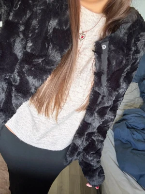 Svart pälsjacka i fuskpäls - Snygg svart pälsjacka i mjuk fuskpäls med kortare modell och diskret knäppning framtill. Jackan har en stilren look och passar perfekt för kalla dagar när du vill vara både varm och trendig.