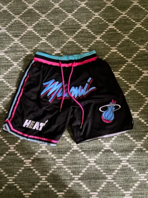 Miami Heat basketshorts svart - Snygga basketshorts i svart med Miami Heat-logga och text i blått, rosa och vitt. Shortsen har elastisk midja med snörning i rosa, samt detaljer i turkos och rosa längs kanterna. Tillverkade i ventilerande meshmaterial, perfekta för basket eller träning.