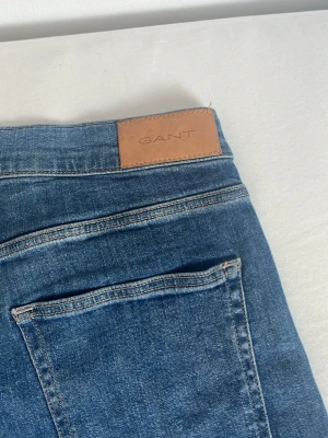Blå jeans från GANT - Snygga blå jeans från GANT med klassisk femficksdesign och läderpatch med logga bak i midjan. Jeansen har en rak passform och är tillverkade i ett mjukt denimtyg som ger en skön känsla. Perfekta för en avslappnad och stilren look. 164 cm , 13-14 år