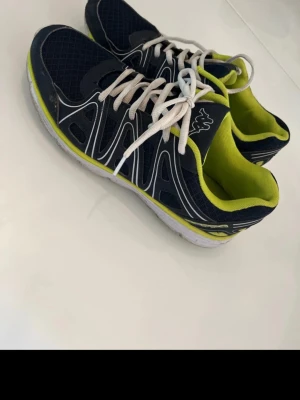 Svarta Kappa sneakers med limegrönt foder - Snygga sneakers från Kappa i svart med vita detaljer och limegrönt foder. Skorna har vita snören, sportig design och meshpartier för bra ventilation. Perfekta för dig som gillar en fräsch och modern look. Yttersulan är vit och ger bra grepp.