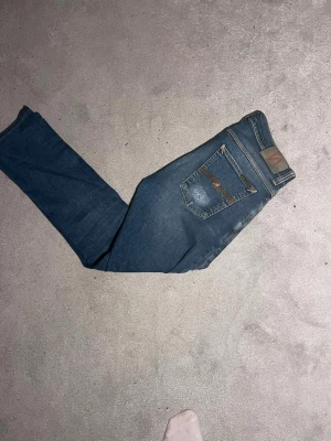 Blå jeans från Nudie Jeans - Säljer ett par klassiska blå jeans från Nudie Jeans med raka ben och snygga slitningar. Jeansen är 41,5 cm i midjan och 100 cm i längd. Modellen på jeansen är tilted tor.