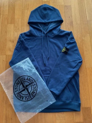 Blå hoodie från Stone Island - Snygg blå hoodie från Stone Island med klassisk patch på vänster ärm. Hoodien har dragsko i huvan och en stor magficka. Tillverkad i mjukt bomullsmaterial som känns skönt mot huden. Är XL men är liten i storlek så har du L så är den perfekt