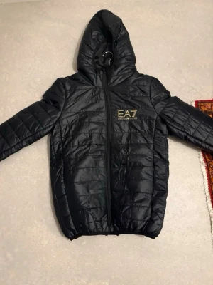 Svart pufferjacka från EA7 Emporio Armani - Snygg svart pufferjacka från EA7 Emporio Armani med huva och guldfärgad logga på bröstet. Jackan har quiltad design, dragkedja framtill och elastiska muddar vid ärmslut och nederkant. Perfekt för kyliga dagar och riktigt trendig look. Utan hängare och lapp inuti 