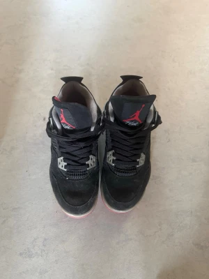 Svarta Air Jordan 4 Retro sneakers - Säljer ett par svarta Air Jordan 4 Retro sneakers med röda och grå detaljer. Skorna har klassisk mesh och mocka på ovandelen, samt den ikoniska Jumpman-loggan på plösen. Insidan är fodrad i rött och sulan har en kontrasterande färg. Perfekta för dig som gillar streetwear.