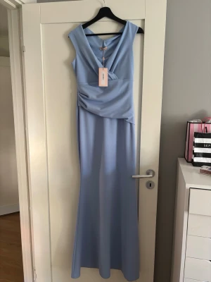 Ljusblå långklänning med v-ringning - Sprillans ny elegant ljusblå långklänning med v-ringad hals och draperad detalj i midjan. Klänningen är ärmlös och har en smickrande, figurnära passform som faller snyggt längs kroppen. Perfekt för speciella tillfällen som en skolbal där du vill känna dig extra fin.