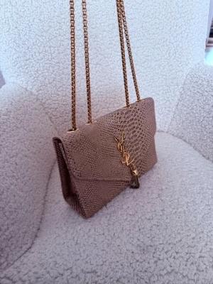 Beige YSL kuvertväska i ormmönster - Säljer en beige kuvertväska från YSL med snyggt ormmönstrat läder, guldfärgad kedja och ikonisk YSL-logga framtill. Väskan har en dekorativ tofs i metall och klassisk kuvertform. Perfekt accessoar för att lyfta din outfit.