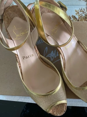 Guldiga sandaler från Christian Louboutin strl 38, kilklack. - Snygga sandaler från Christian Louboutin i guld med öppen tå och justerbar rem runt ankeln. Ovandelen har ett metalnät och övrig material i guldfärg, sulan är röd i äkta läder. Perfekta för dig som vill sticka ut med exklusiva detaljer och lyxig känsla. Obs! Liten i storleken och passar inte bredda fötter.