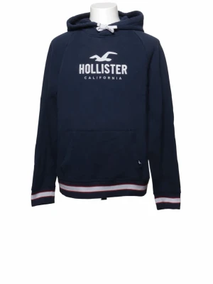 Mörkblå hoodie från Hollister - Mörkblå hoodie från Hollister med vit logotyp och text på bröstet. Den har huva med snören, en stor magficka och ribbade muddar med vita och röda ränder. Perfekt för dig som gillar en avslappnad stil.