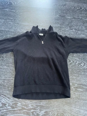 Svart stickad halvzip tröja - Säljer en svart stickad tröja med halvzip och hög krage. Tröjan har ribbade muddar vid ärmslut och nederkant samt en grovstickad struktur. Perfekt för lager på lager och enkel att styla till olika outfits.