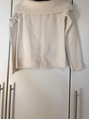 Vit stickad tröja från Gina Tricot - Mysig vit stickad tröja med bred, ribbad krage som kan vikas ner. Tröjan har långa ärmar och ribbade muddar vid ärmslut och nederkant. Perfekt för kyliga dagar och enkel att matcha med olika outfits.
