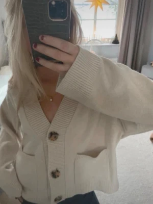 Beige oversized kofta med stora knappar - ❗️inte min bild❗️Säljer en beige oversized kofta med djup V-ringning och stora, mönstrade knappar framtill. Koftan har breda ribbade muddar och två stora fickor framtill. Perfekt för lager på lager och en avslappnad look.