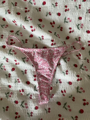 Rosa stringtrosa med blommigt mönster - Supersöt bikiniunderdel i stringmodell med rosa blommigt mönster på vit botten. 