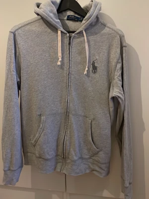 Grå zip hoodie från Polo Ralph Lauren - Klassisk grå zip hoodie från Polo Ralph Lauren med huva och vita dragsnören och tunnare tröja . Broderad logga i mörkgrått på bröstet och två stora fickor framtill. Tillverkad i mjuk bomullsblandning för en skön och avslappnad look.
