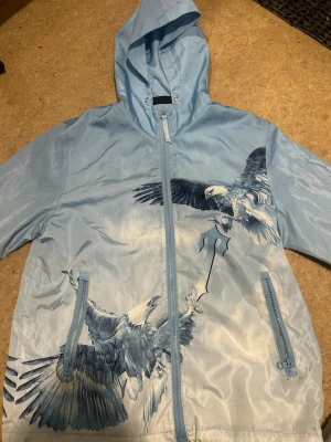 Trapstar Windbreaker - Snygg ljusblå vindjacka från Trapstar med stort tryck av två örnar på både fram- och baksida. Jackan har huva, dragkedja framtill och två fickor med dragkedja. Materialet är glansigt och lätt, perfekt för streetwear-stil. Trapstar-loggan syns tydligt på ryggen.