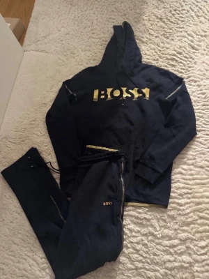 Svarta/mörkblått mjukisset från BOSS - Snygga svarta/mörkblåa mjukisbyxor från BOSS med gulddetaljer och logga på benet, de har en matchande överdel till. Byxorna har dragsko i midjan och guldfärgade ränder längs sidorna. Perfekt för en chill och trendig look.