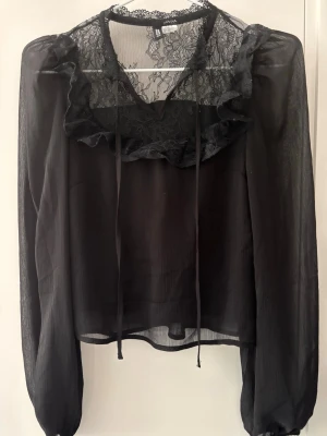 Svart blus med spets och volang - Snygg svart blus från HM med transparenta ärmar, spetsdetaljer vid halsen och volang framtill. Blusen har knytband vid halsen och en lätt, luftig känsla. Perfekt för dig som gillar trendiga och feminina plagg.