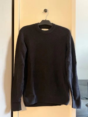 Mörkblå stickad tröja från Casual Friday - Säljer en mörkblå stickad tröja med snyggt zigzagmönster över hela plagget. Tröjan har rund halsringning, långa ärmar och ribbade muddar vid ärmslut och nederkant. Perfekt för dig som gillar stilrena detaljer och en klassisk look.