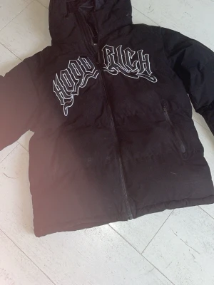 Svart pufferjacka från Hoodrich - Svart pufferjacka från Hoodrich med stor vit logga på bröstet. Drag kedjan sönder går att fixa hos skräddare Jackan har dragkedja framtill, två sidofickor med dragkedja och en hög krage med huva. Perfekt för kalla dagar och ger en streetwear vibe.