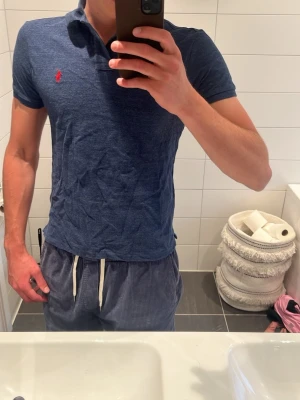 Blå pikétröja från Ralph Lauren - Snygg marinblå pikétröja från Ralph Lauren med klassisk krage och två knappar framtill. Tröjan har en röd broderad logga på bröstet och är i en slim passform. Perfekt för en stilren och avslappnad look. Modellen är 187cm och väger 80 kg, hör av er om prisförslag!!