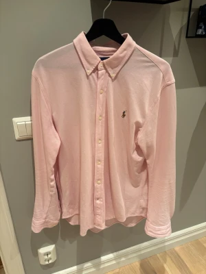 Ljusrosa skjorta från Ralph Lauren - Klassisk ljusrosa skjorta från Ralph Lauren med broderad logga på bröstet. Skjortan har knappar hela vägen, krage och långa ärmar. Sparsamt använd!