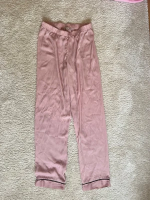 Rosa pyjamasbyxor från Anna Field - Mysiga pyjamasbyxor i ljusrosa från Anna Field. Byxorna har resår i midjan och är gjorda i ett mjukt, glansigt material. Nedtill finns en tunn svart kant som detalj. Perfekta för chillkvällar hemma.
