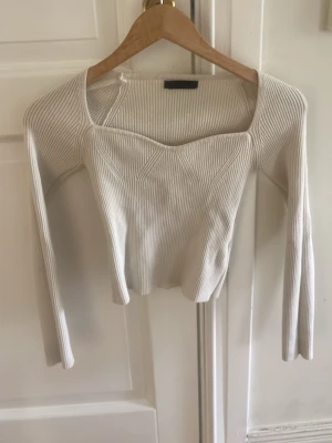 Vit ribbad långärmad topp - Snygg vit ribbad topp med lång ärm och unik skärning vid halsen. Toppen har en croppad passform och är tillverkad i ett stretchigt material som sitter tajt mot kroppen. Perfekt att styla med jeans eller kjol för en trendig look.