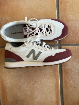 New Balance vita och vinröda sneakers - Snygga sneakers från New Balance i vitt och vinrött med grå detaljer och klassisk N-logga på sidan. Ovandelen är i mesh och mocka, med vita skosnören och en bekväm sula. Perfekta för en sportig och avslappnad stil.