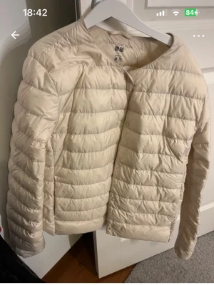 Beige ultralätt dunjacka Uniqlo - Snygg beige dunjacka från Uniqlo i ultralätt material. Jackan har en minimalistisk design utan krage, horisontella sömmar och tryckknappar framtill. Perfekt för kalla vinterdagar och enkel att bära under andra plagg.