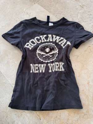 Svart t-shirt Rockaway New York - Svart t-shirt med tryck 'Rockaway New York' och basebollmotiv på bröstet. Klassisk rund hals och korta ärmar. 