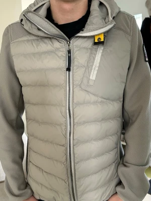 Ljusgrå pufferjacka från Parajumpers - Superfräch och snygg ljusgrå jacka från Parajumpers med huva och dragkedja framtill. Jackan har quiltad front och rygg, samt släta ärmar med patch på axeln. Perfekt för kyliga dagar och har en sportig vibe. Hel och inga defekter och självklart äkta! 