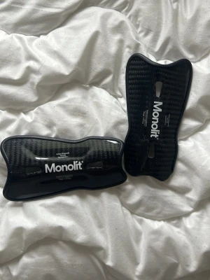 Monolit benskydd i kolfiber - Säljer ett par svarta benskydd från Monolit i kolfiber-look. De är lätta och väger 30 gram, smidiga och har en modern design med vit logga. Perfekta för dig som vill ha extra skydd och stil på fotbollsplanen.