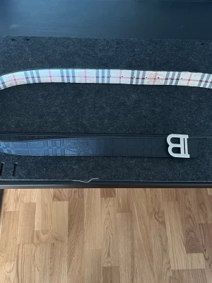 Reversible Belt Burberry - This reversible belt features a classic beige, red, and black check pattern on one side, reminiscent of iconic designer styles. The reverse side is black with a distinct embossed texture, possibly crocodile-effect, and is adorned with a prominent silver-toned 'B' buckle.   #reversiblebelt #Burberry#buckle #texturedleather #fashionaccessory #mensbelt #womensbelt #style #secondhand #preloved