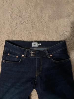 Mörkblå bootcut jeans från 157 - Mörkblå jeans från 157 med klassisk femficksmodell och bootcut-ben. Jeansen har kontrastsömmar i gult och dubbla knappar i midjan. Perfekta för dig som gillar en tidlös och stilren look. Storlek M short 