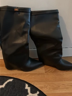 Svarta höga stövlar i skinnimitation - Säljer ett par svarta höga stövlar med spetsig tå och bredare skaft. Stövlarna är tillverkade i skinnimitation och har en enkel, stilren design utan synliga detaljer förutom en liten metallogo upptill. Perfekta för att lyfta din outfit.