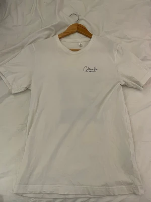 Vit t-shirt från Culture de la mode - Vit t-shirt med tryck från Culture de la mode. Framsidan har en liten logga på bröstet och baksidan har ett större svartvitt fototryck med text. Klassisk passform och rund halsringning. Perfekt för en clean och stilren look.