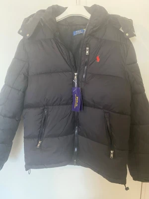 Svart dunjacka från Polo Ralph Lauren - Svart dunjacka från Polo Ralph Lauren med röd broderad logga på bröstet. Jackan har huva, flera dragkedjefickor och quiltad design. Perfekt för kalla vinterdagar och riktigt snygg streetstil.