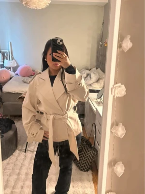 Beige kort trenchcoat med bälte - Trendig beige trenchcoat. Perfekt för lager-på-lager och snygg till jeans eller kjol. Har aldrig använt. Nypris:1099