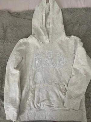 Ljusgrå hoodie från GAP - Ljusgrå hoodie från GAP med broderad logga framtill. Klassisk modell med stor huva och känguruficka. Mjuk bomullsblandning som är perfekt för en avslappnad stil. Snygg och enkel att matcha med jeans eller mjukisbyxor.