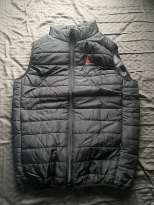 Ralph Lauren väst - Snygg svart dunväst från Ralph Lauren med klassisk  design och röd logga på bröstet. Västen har hög krage, dragkedja framtill och två fickor. Perfekt för lager-på-lager och enkel att matcha med olika outfits. 