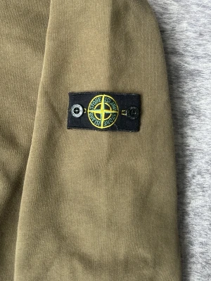 Olivgrön hoodie från Stone Island - Snygg olivgrön hoodie från Stone Island Junior med klassisk logotyp-patch på ärmen. Tillverkad i mjuk bomull med ribbade muddar vid hals och ärmslut. Perfekt för dig som gillar streetwear och vill ha en stilren look. XXS men passar väldigt bra som XS