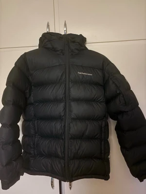 Svart dunjacka från Peak Performance - Snygg svart dunjacka från Peak Performance med hög krage och dragkedja framtill. Jackan har en puffig design och diskret logga på bröstet. Perfekt för kalla dagar när du vill hålla dig varm och stilren.(nypris 4199kr) priset kan diskuteras.
